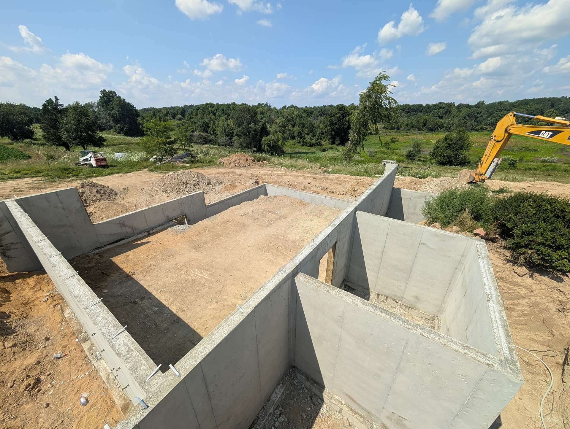 A concrete house foundation under construction on a sunny day with an excavator visible in the background.