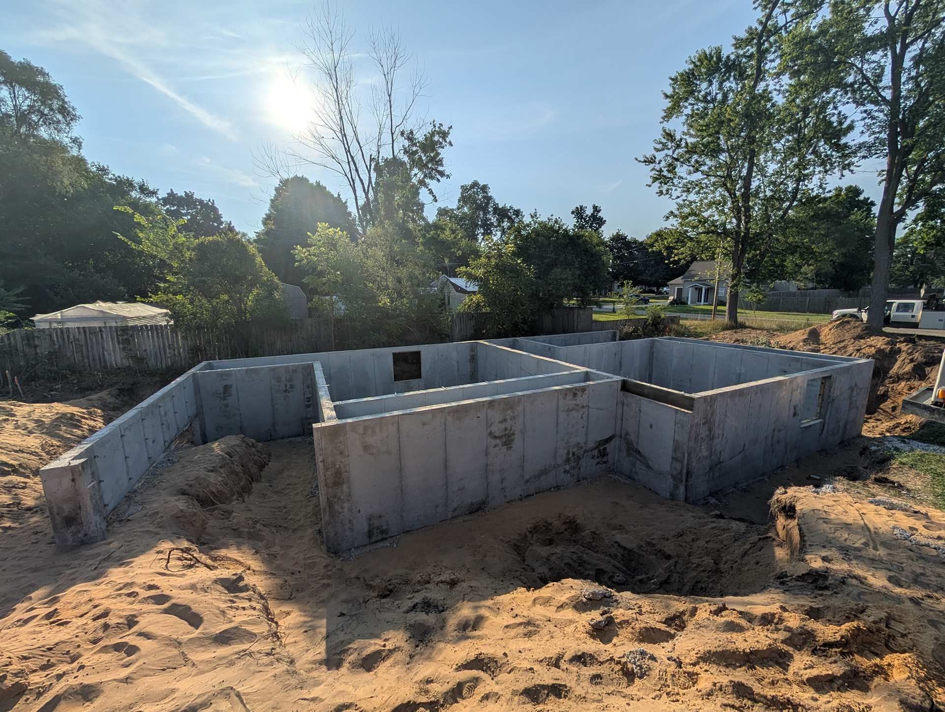 A concrete foundation for a house under construction, surrounded by dirt, under a bright sun and trees.