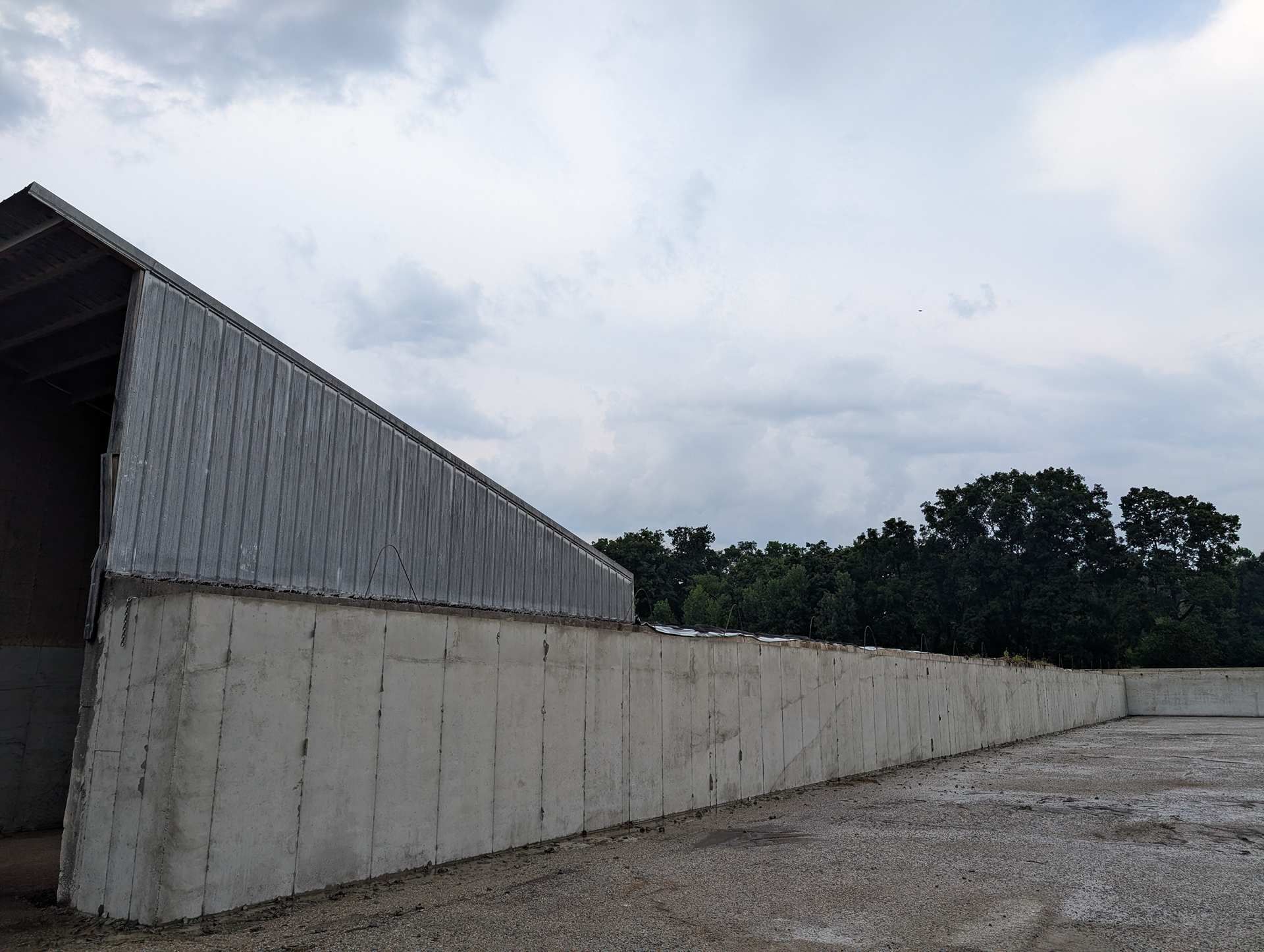 A long concrete retaining wall supporting a metal roof structure, bordering an empty outdoor lot under a cloudy sky.