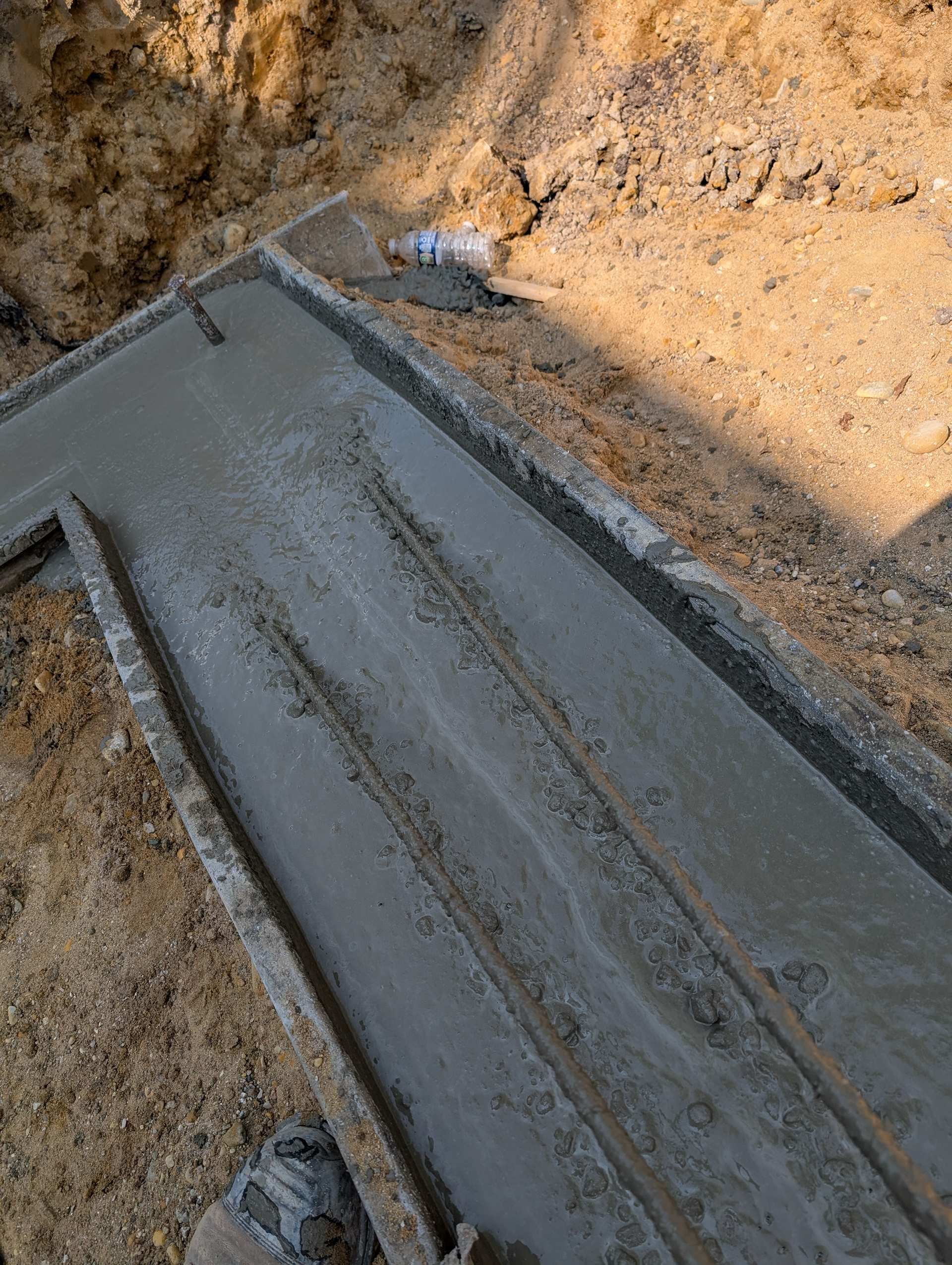 A top-down view of a freshly poured concrete trench footing set in a excavated dirt foundation.