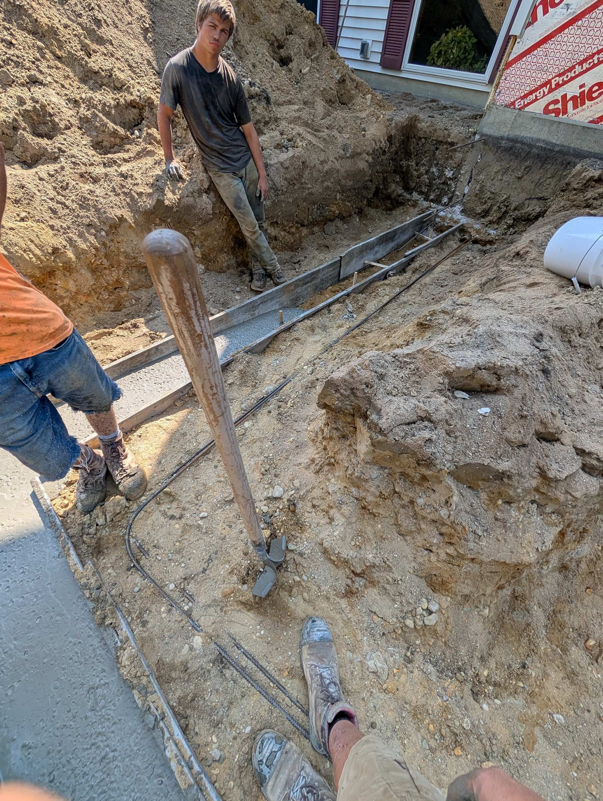 Two people work in a trench next to a building foundation, with a shovel standing upright in the dirt.