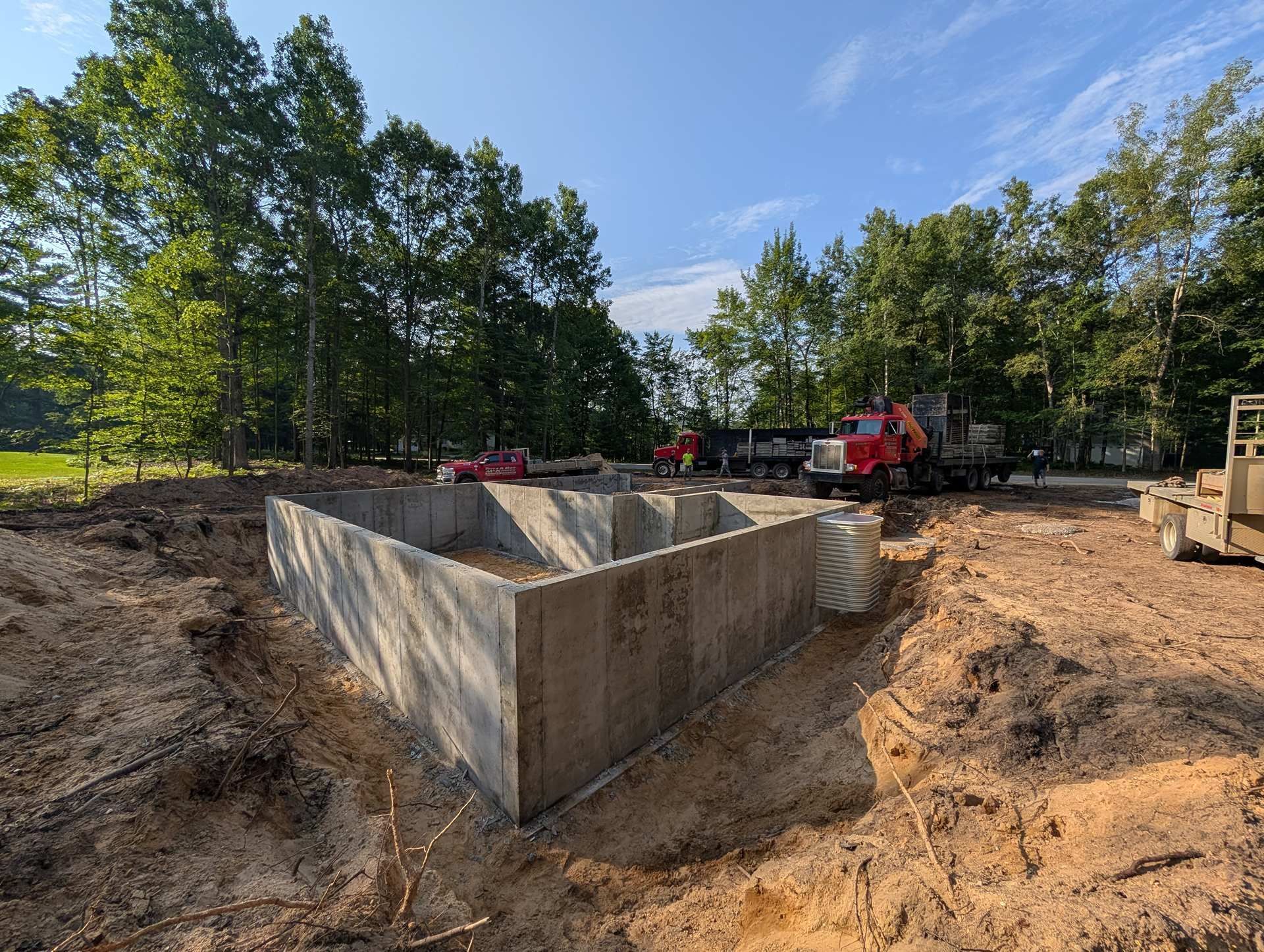 Concrete foundation walls under construction at a wooded job site with dump trucks parked in the background.