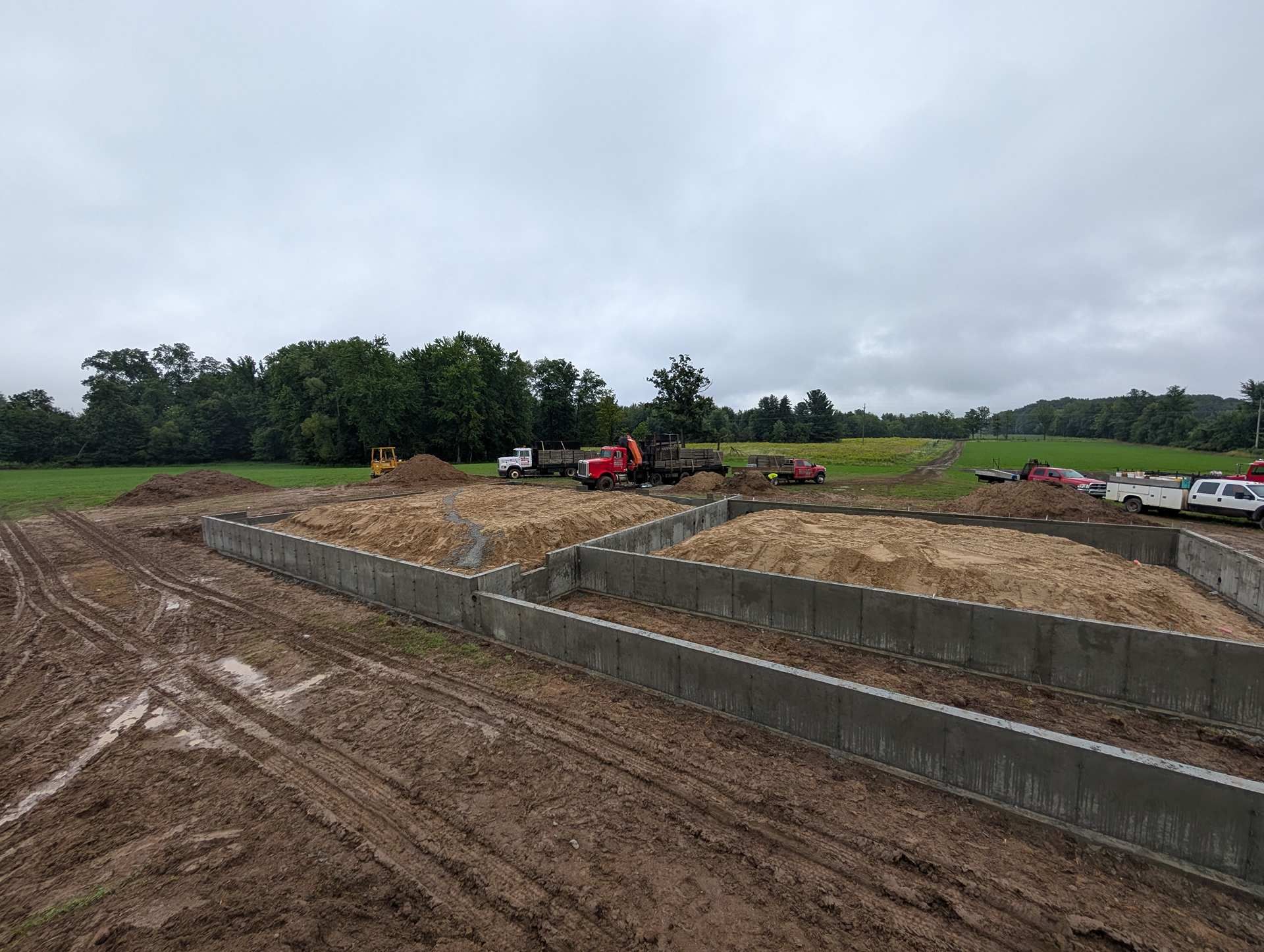 A construction site showing concrete foundation walls filled with soil, with heavy machinery and vehicles in the distance.