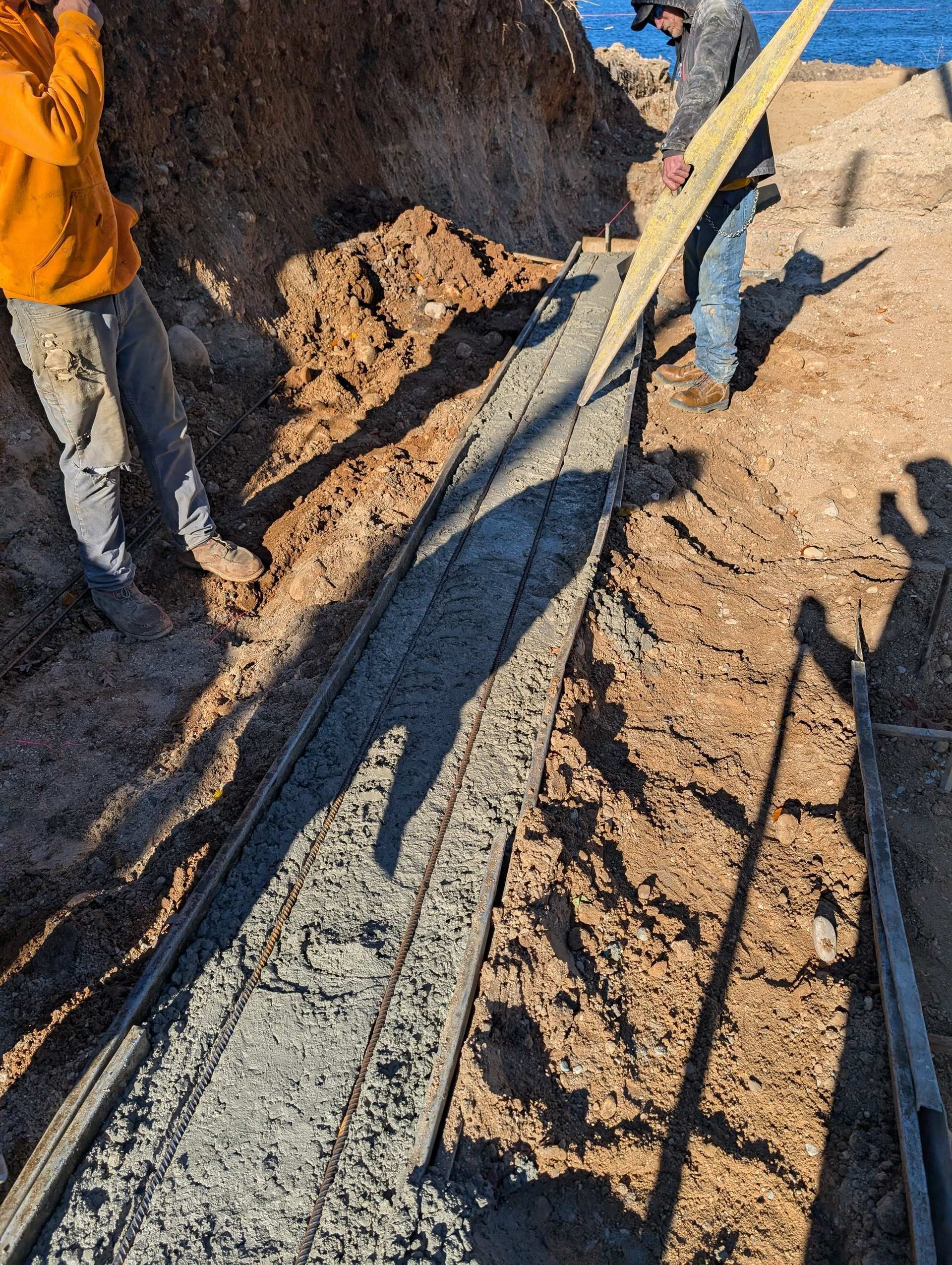 Two workers standing in an excavated trench, one leveling fresh concrete between wooden forms.