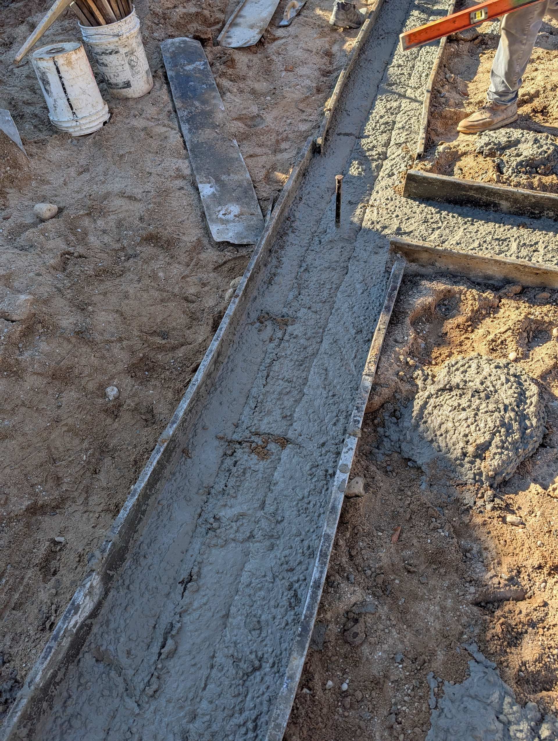 Freshly poured concrete in narrow trench forms on a sandy construction site with nearby buckets and tools.