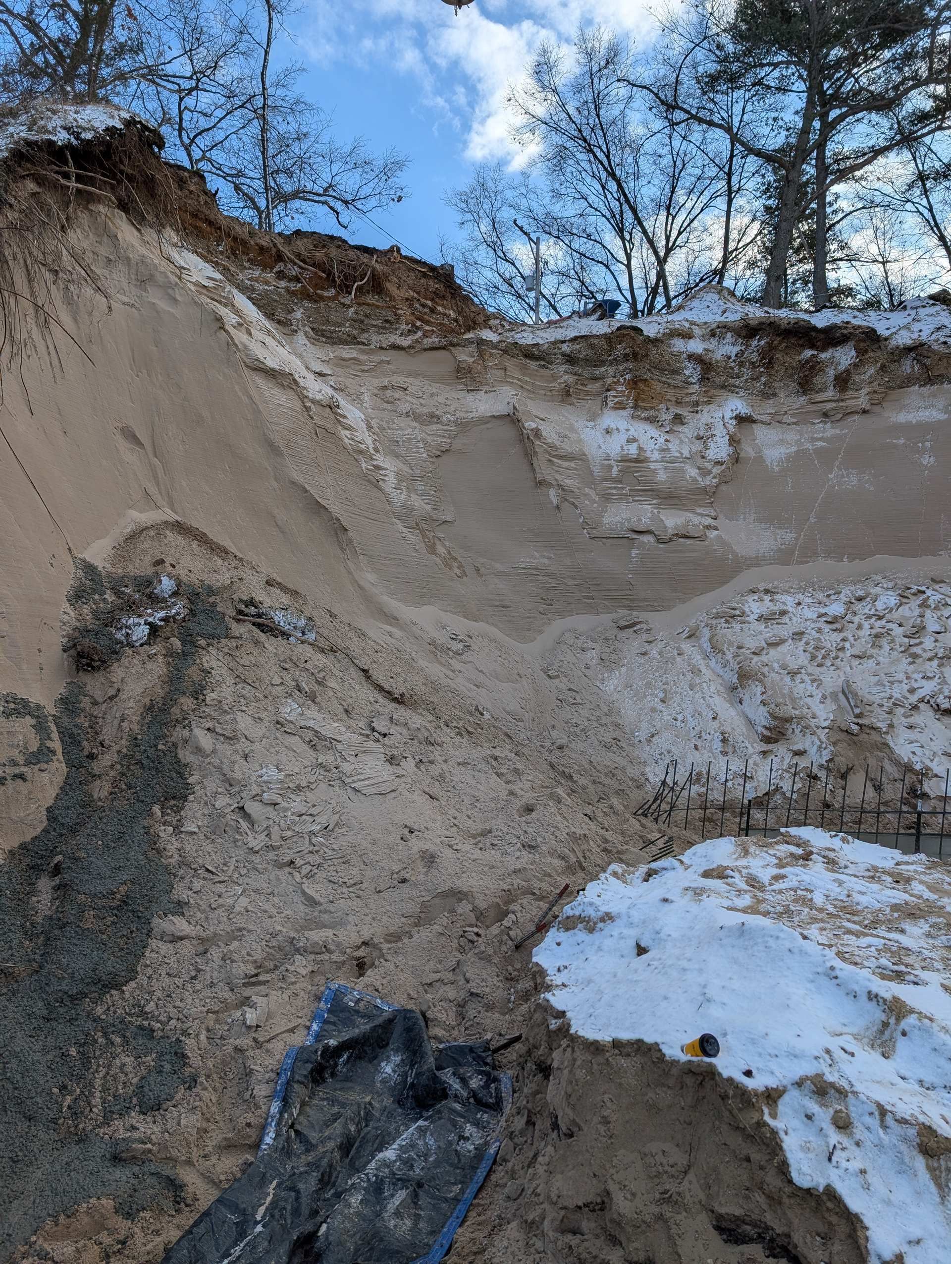 A steep, sandy embankment with exposed soil layers partially covered by patches of snow under a cloudy blue sky.