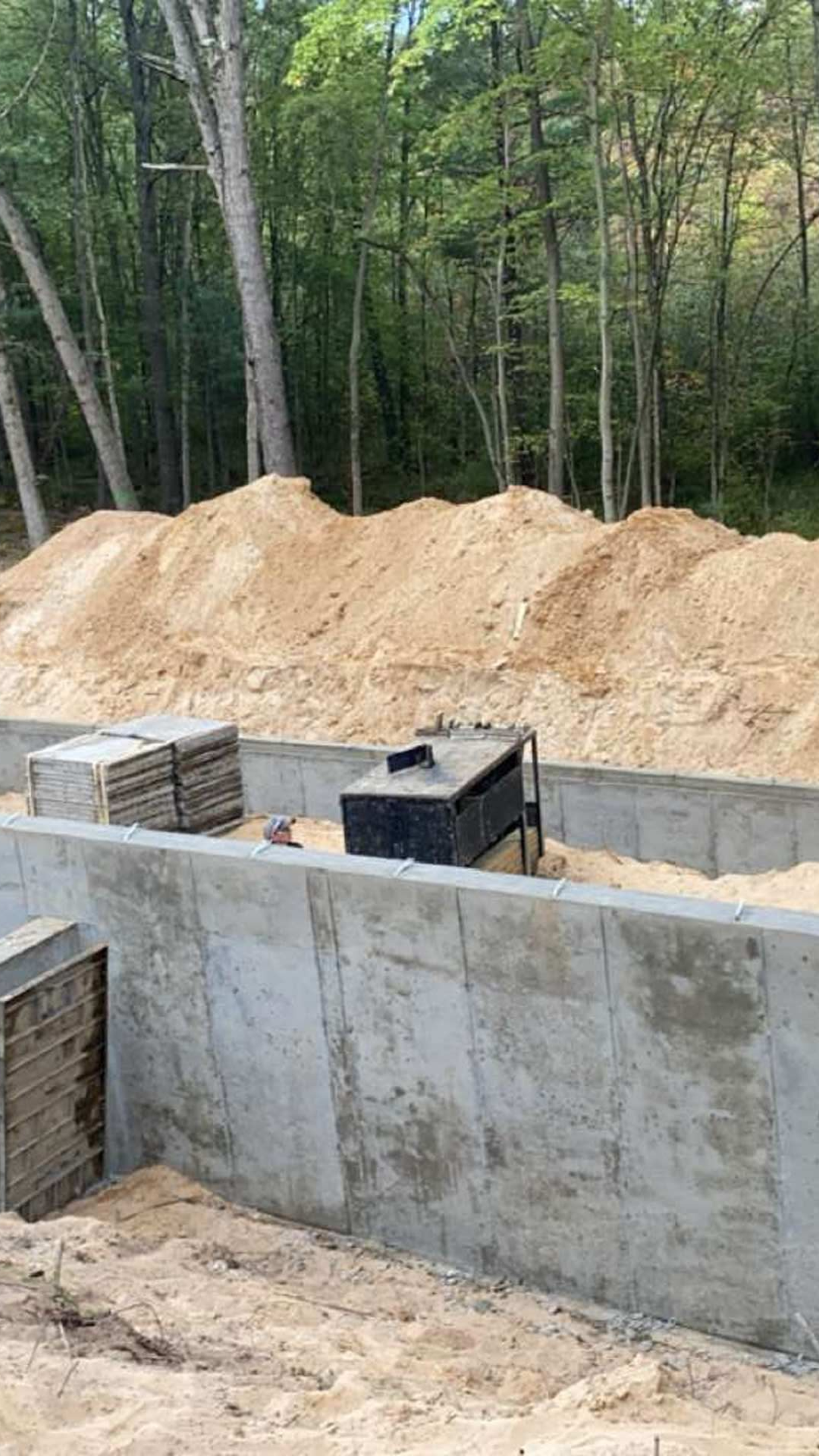 A construction site with gray concrete foundation walls, stacks of blocks, and piles of sand against a backdrop of trees.