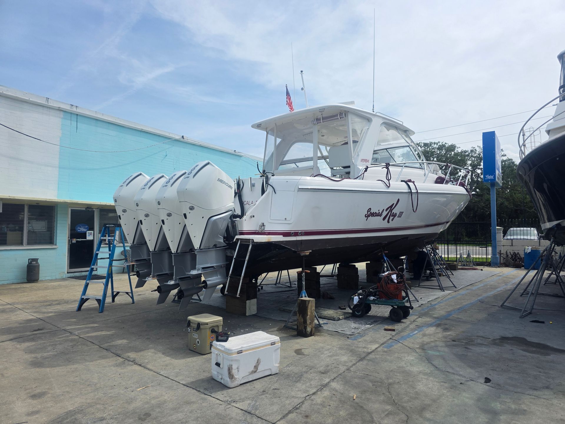 White boat on dry dock with covered hull sections, parked beside a light-blue building under a clear sky.