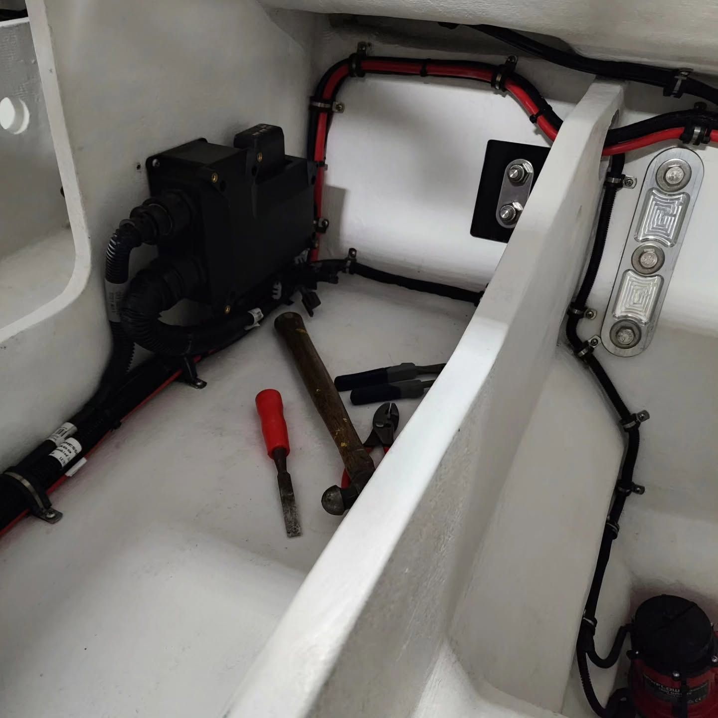 White boat compartment with wiring, hoses, a black pump, and red/black tools on a molded fiberglass floor.