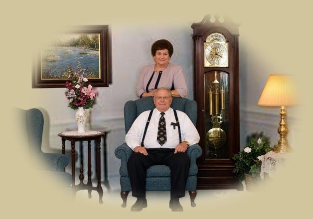 Man seated in teal chair with woman standing behind; grandfather clock, lamp, flowers, and landscape painting nearby.