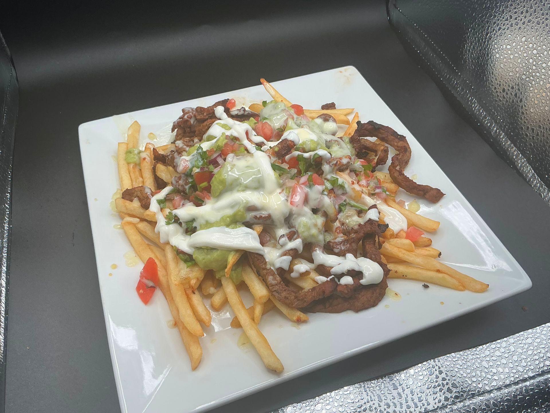 Fries topped with meat, guacamole, salsa, and cream on a white square plate.