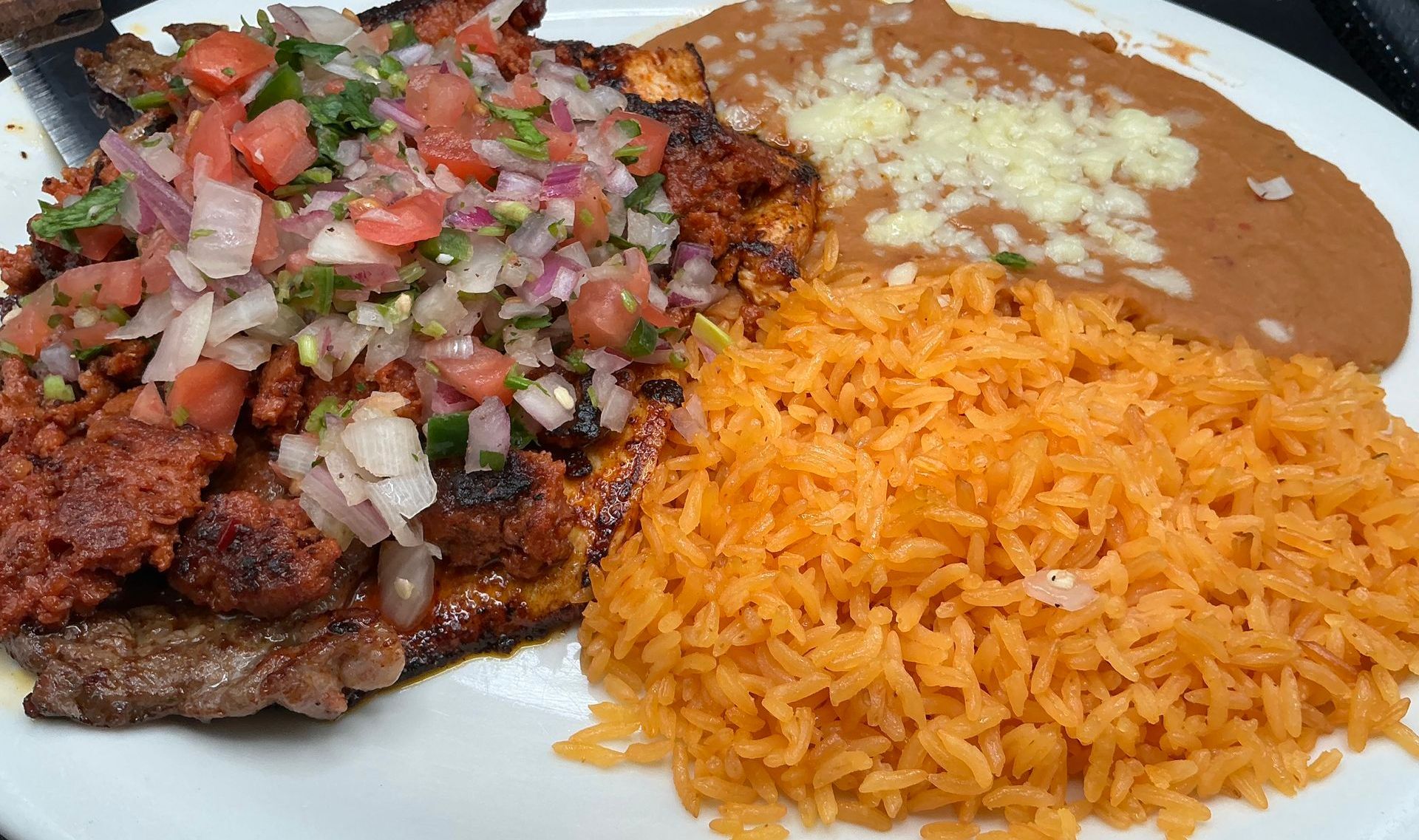 Grilled meat topped with salsa, rice, and refried beans on a white plate.
