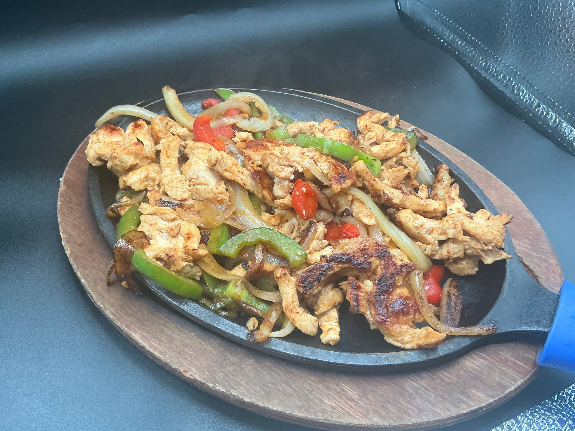Sizzling chicken fajitas with onions and peppers on a hot cast iron plate, wooden base.