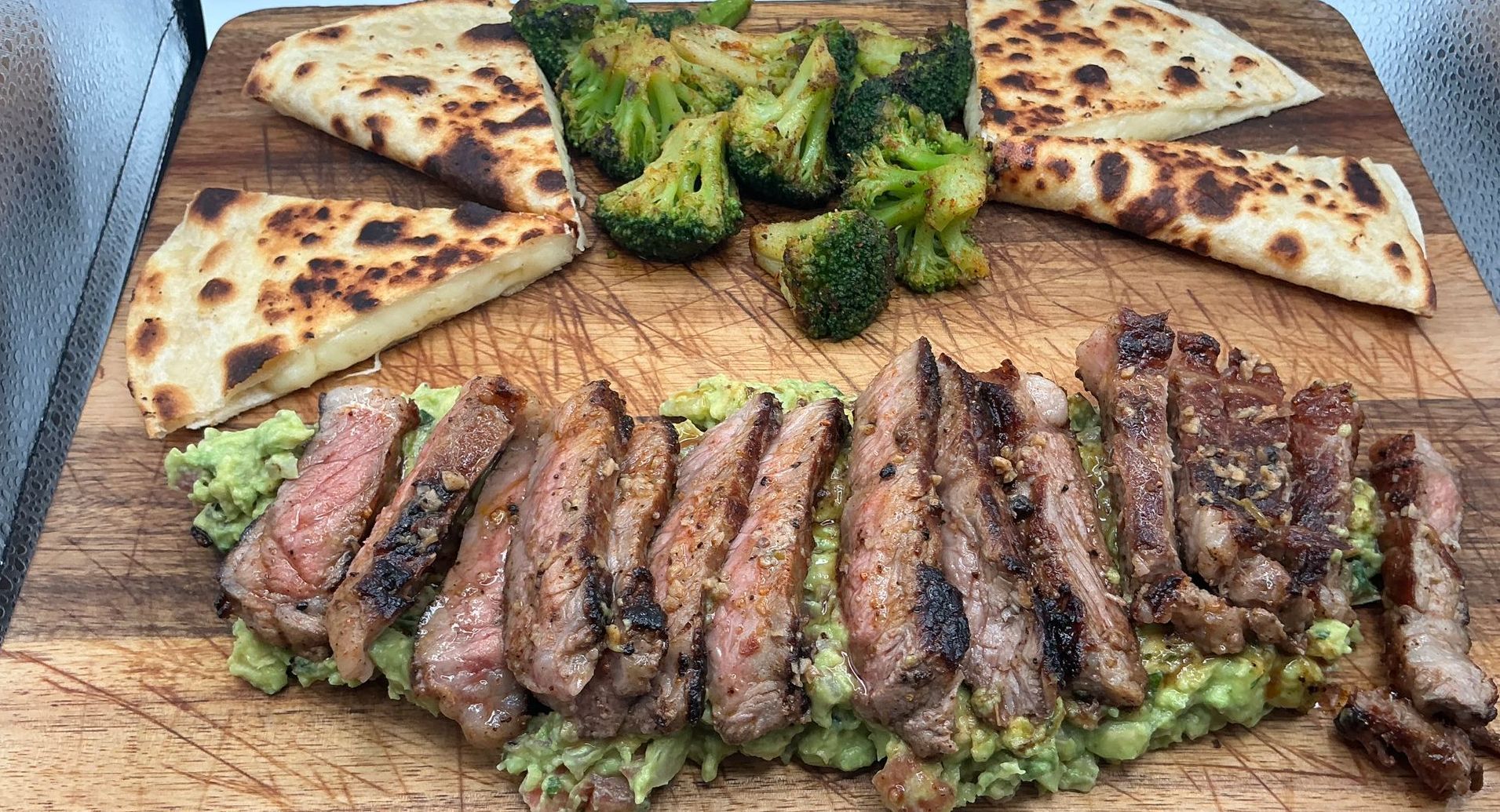 Grilled steak slices over guacamole, with broccoli and naan bread on a wooden board.