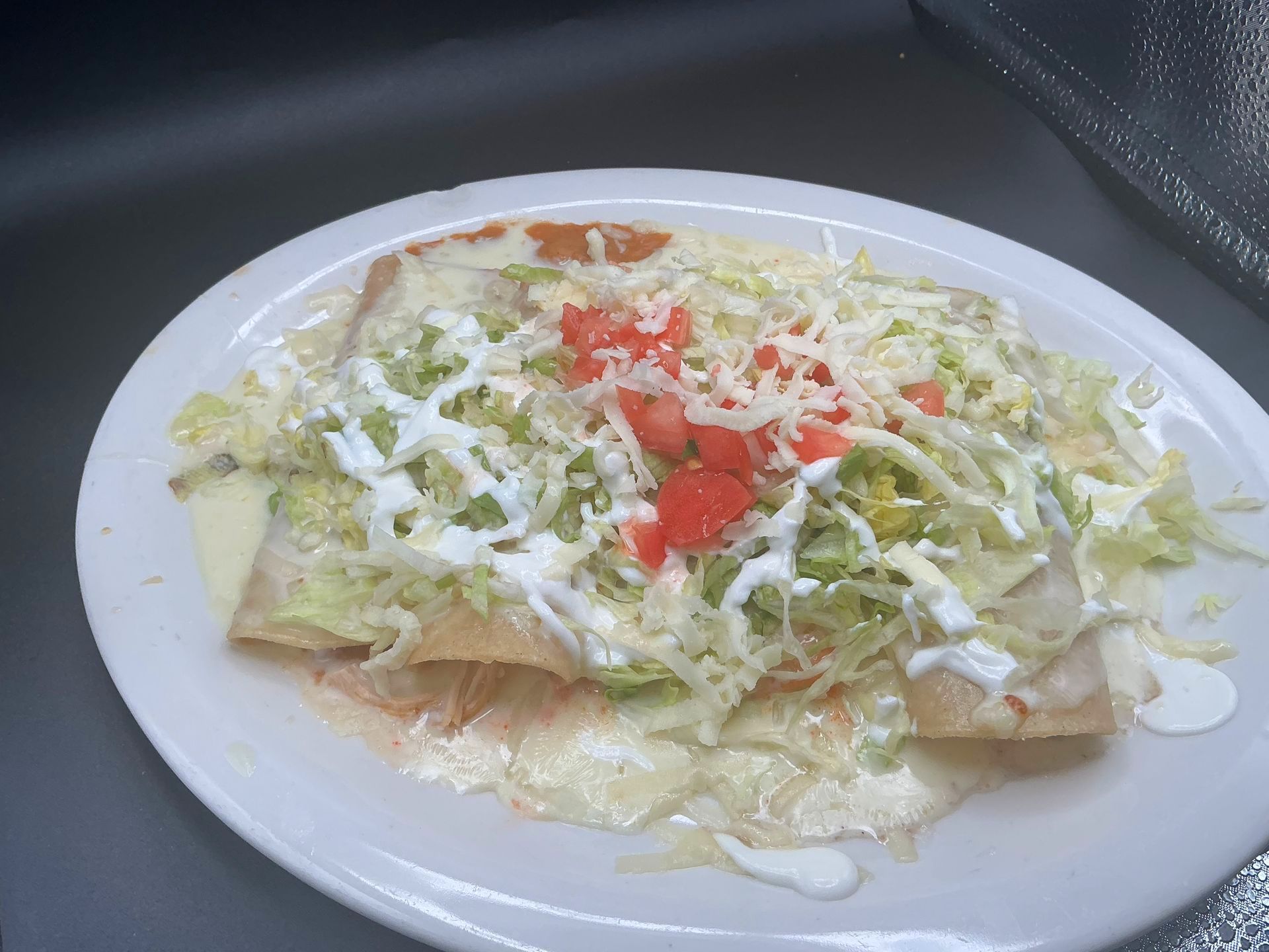 Enchiladas on a white plate, topped with shredded lettuce, cheese, tomatoes, and drizzle of white sauce.