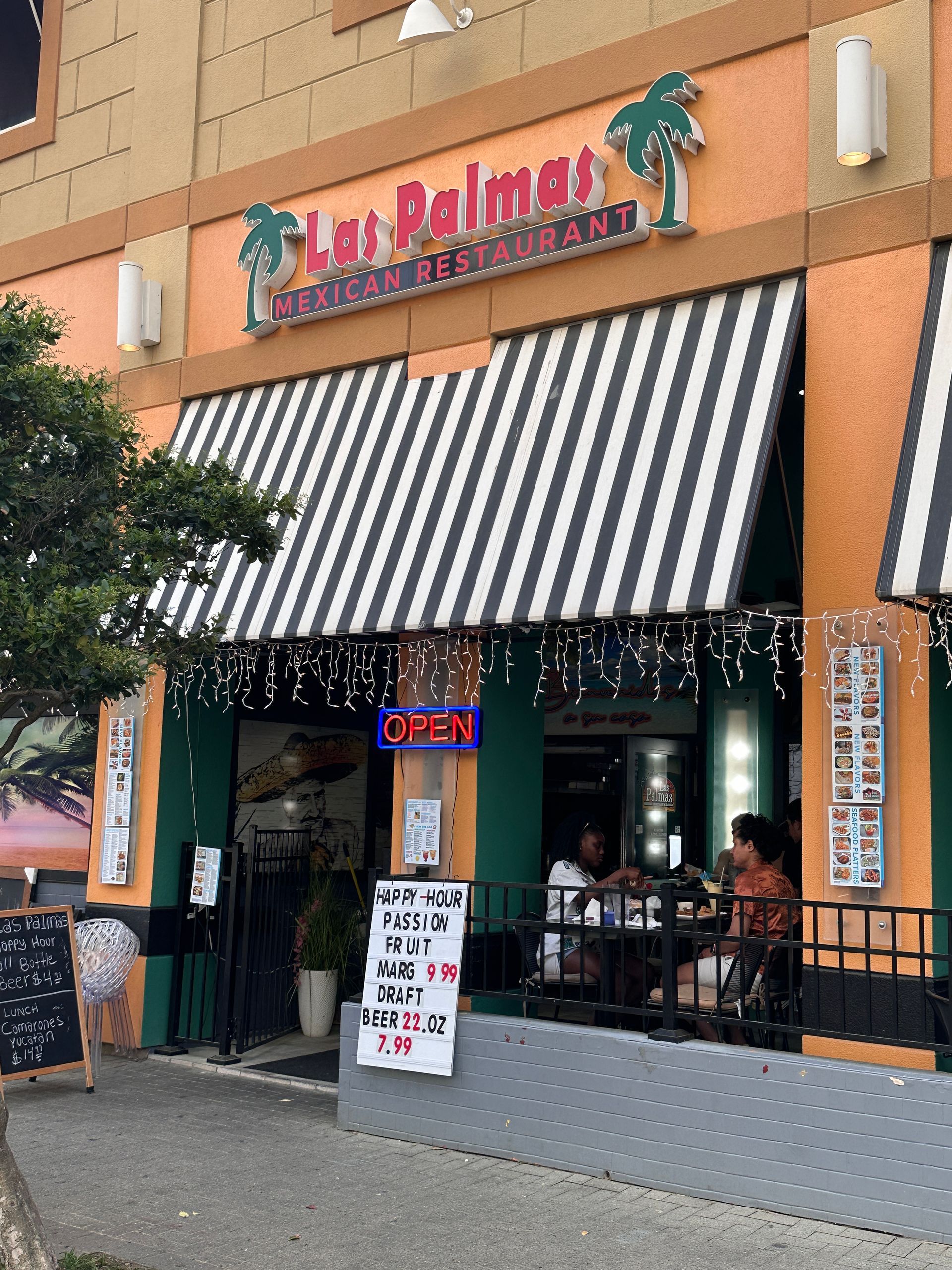Restaurant exterior with striped awning and palm tree sign. People sit at outdoor tables.