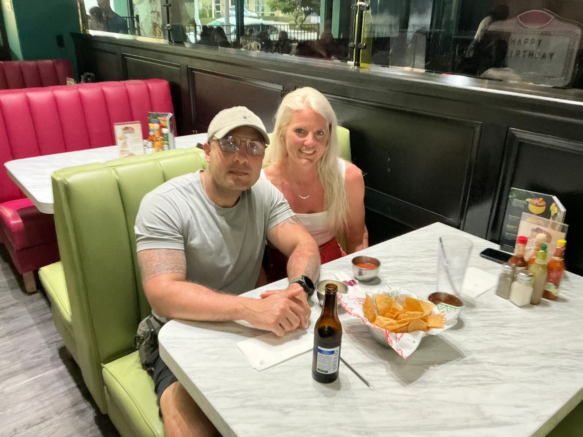 Couple sitting at a table in a diner, with food and a beer. Green and pink booths are visible.