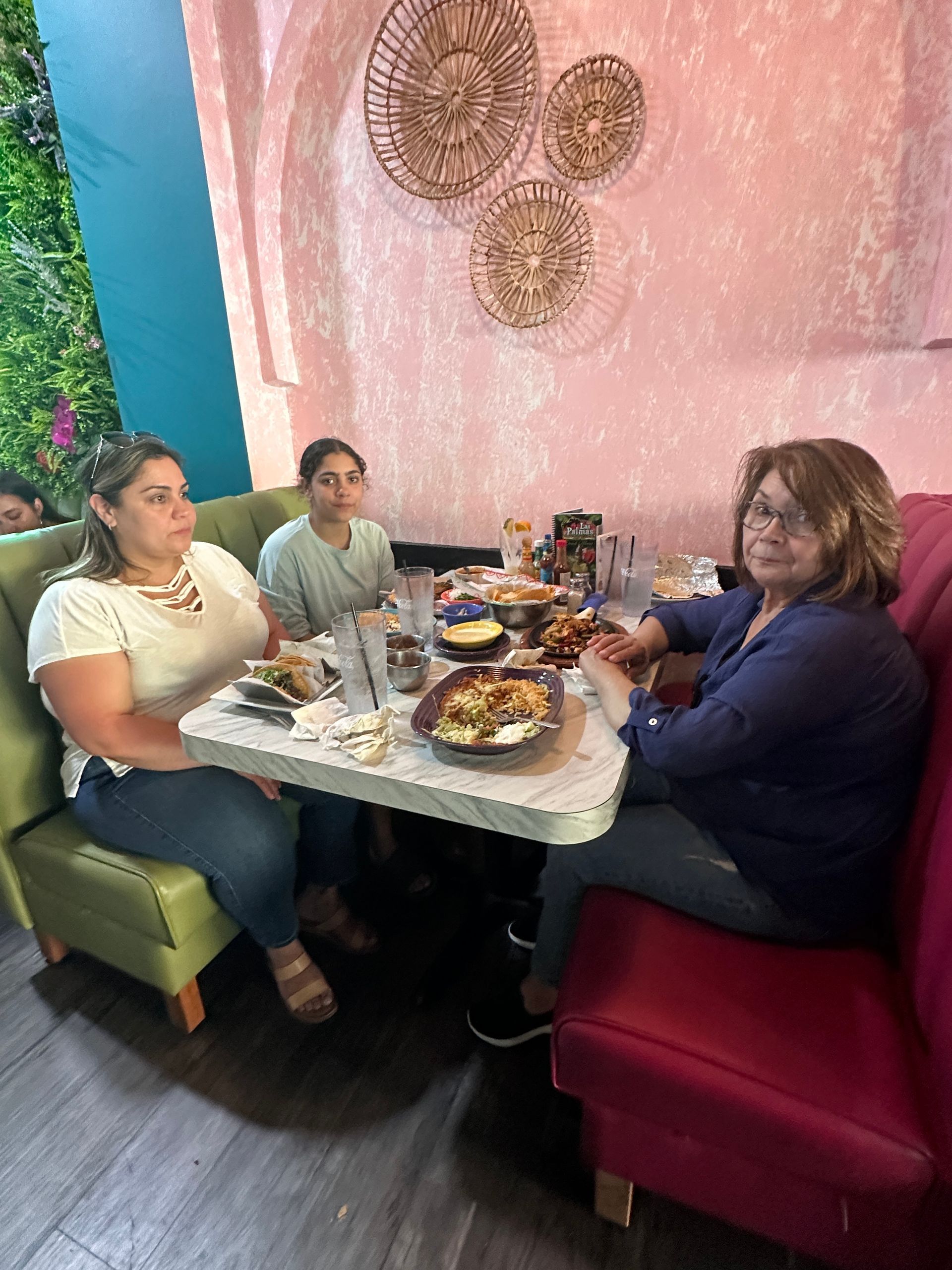 Three people seated at a restaurant table with food, pink walls, and decorative wall hangings.