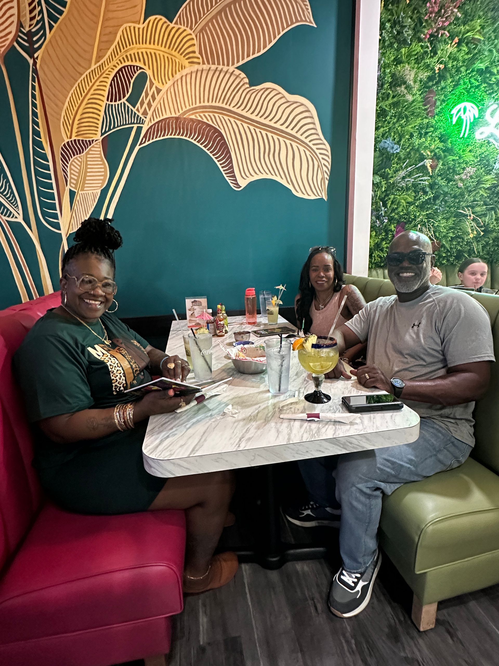 Three people at a restaurant table. Two women and a man are smiling. Table has drinks and food. Teal and gold wall decor.