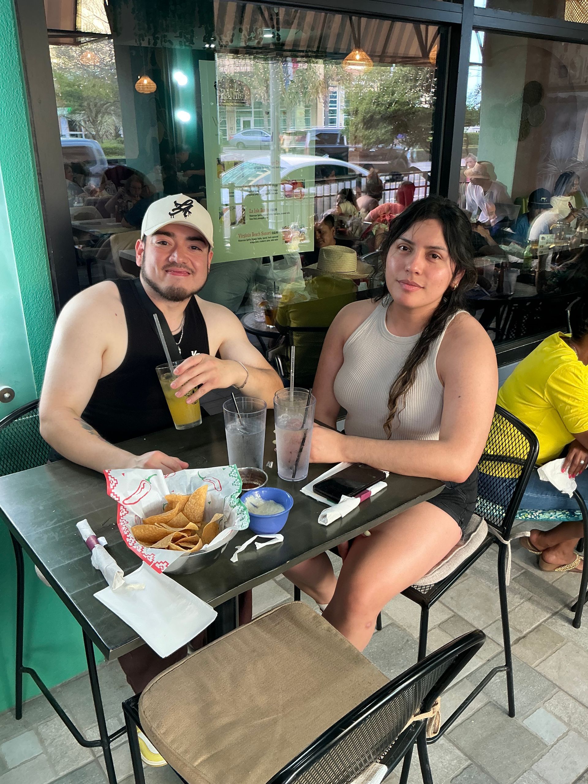Two people seated at a restaurant table with drinks and chips. A window is in the background.