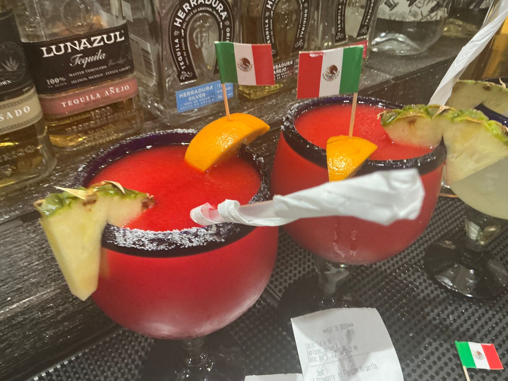 Two red cocktails garnished with orange slices and pineapple, Mexican flags, and tequila bottles.