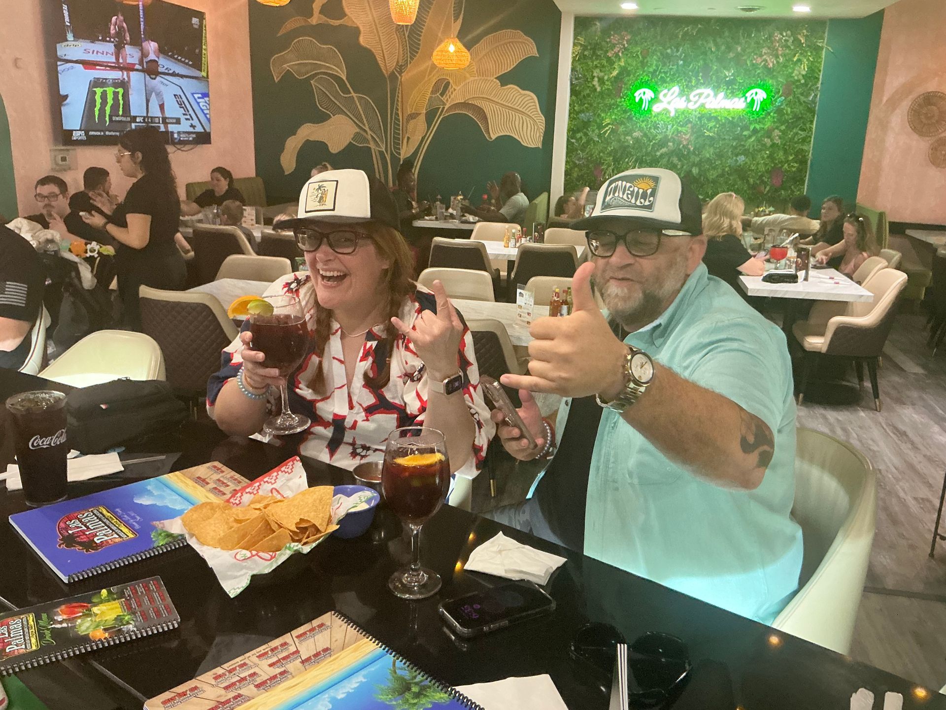 Two people at a restaurant, smiling and gesturing with drinks. Tables and other diners are visible.