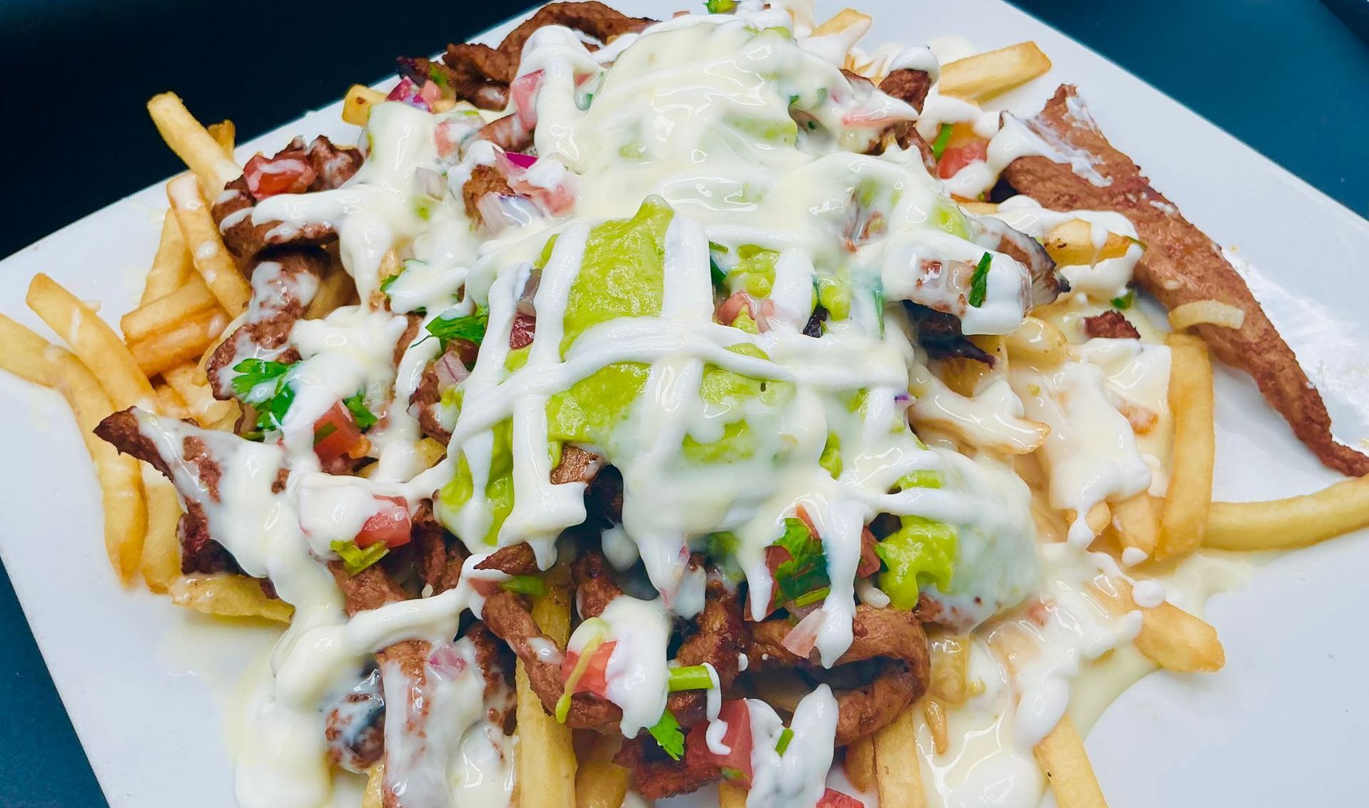 Steak fries topped with guacamole, pico de gallo, and white sauce on a white plate.