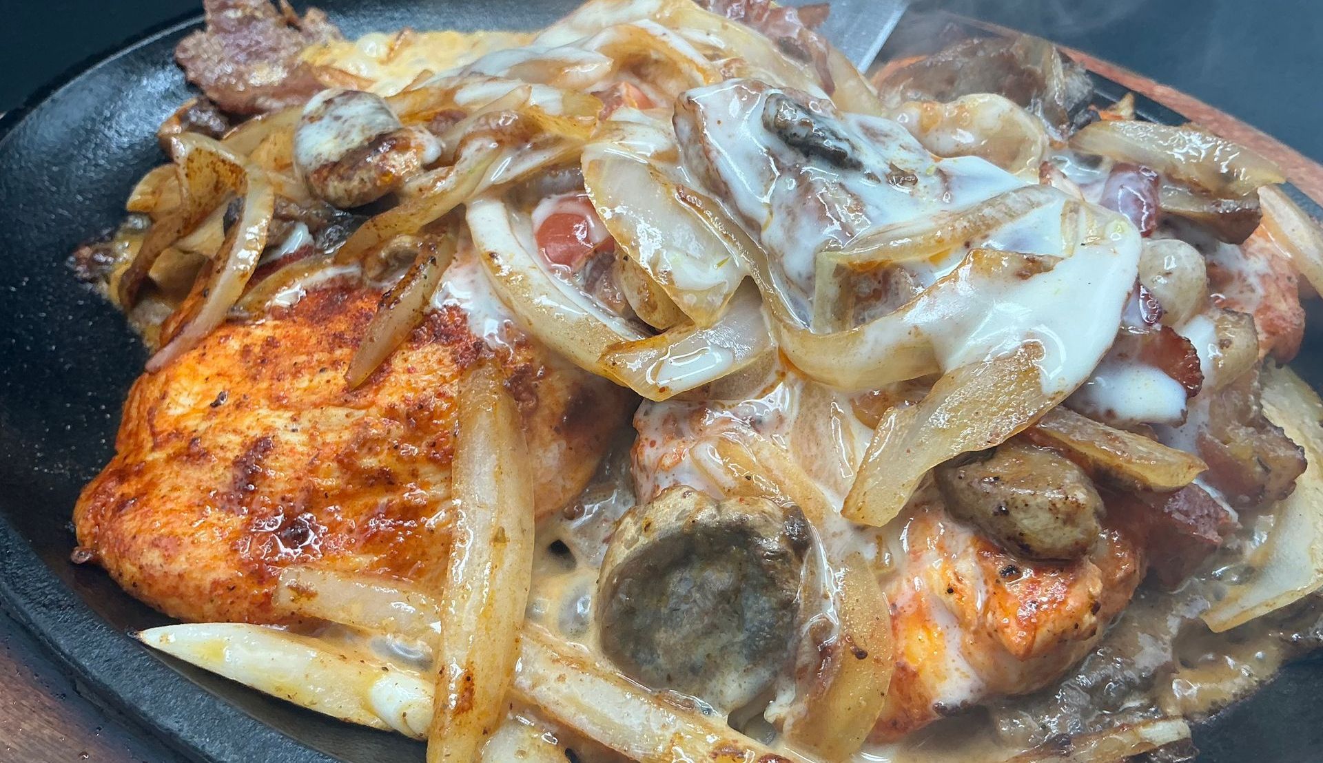 Sizzling plate of food with chicken, onions, mushrooms, and sauce, possibly fajitas, on a dark background.