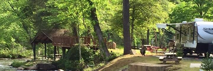 Wooded park with a small pavilion, picnic table, creek, and an RV parked beside a road