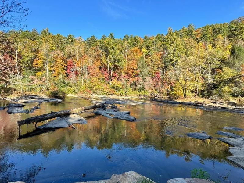 Rocky river with calm reflections, framed by colorful autumn trees under a clear blue sky