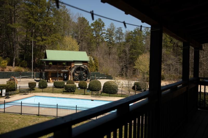 Cabin with green roof beside a pool, viewed from a shaded porch in a wooded area