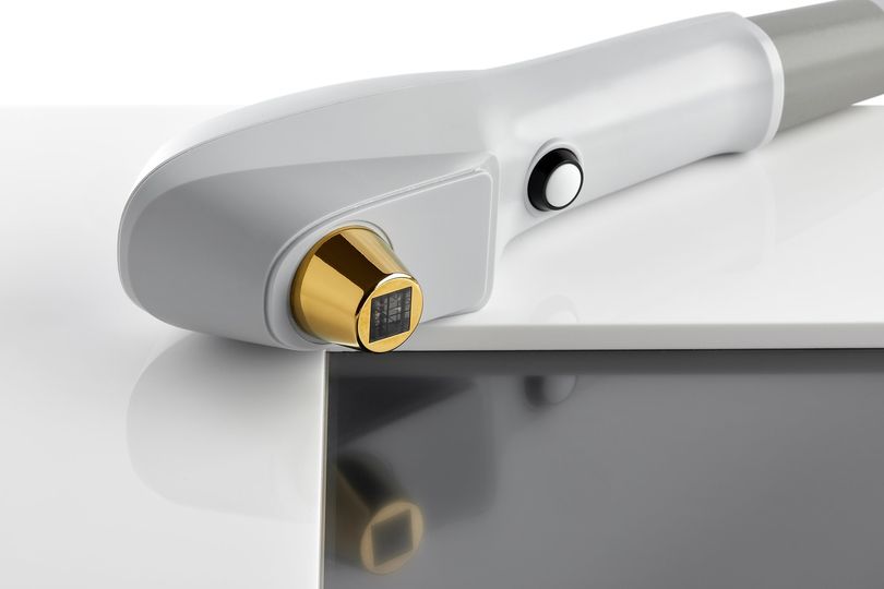 Manipolo per la depilazione laser, grigio e oro, appoggiato su una superficie bianca, con riflesso.