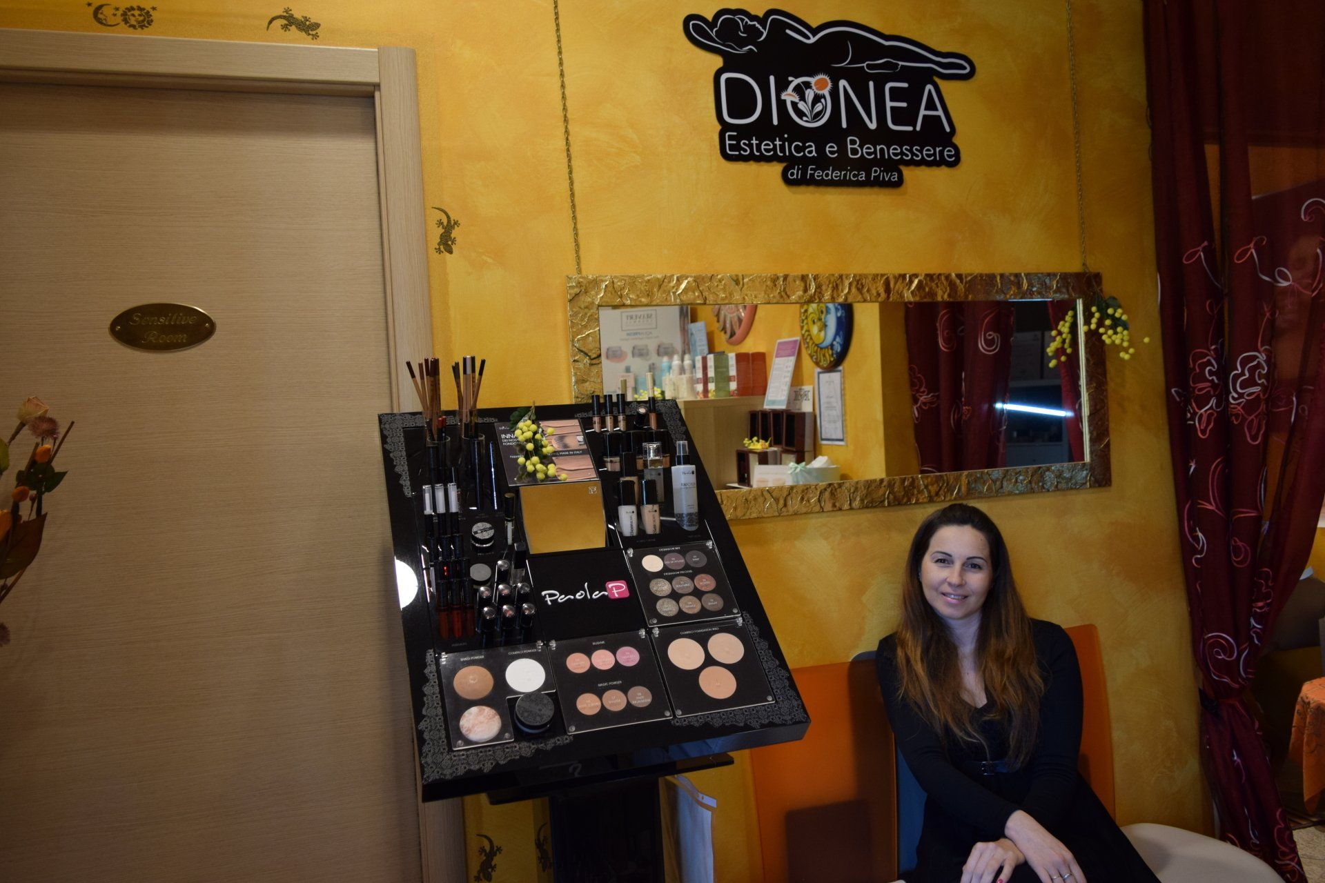 Donna con blazer nero seduta vicino a un espositore per trucchi in un salone di bellezza, accanto a uno specchio e a un cartello.