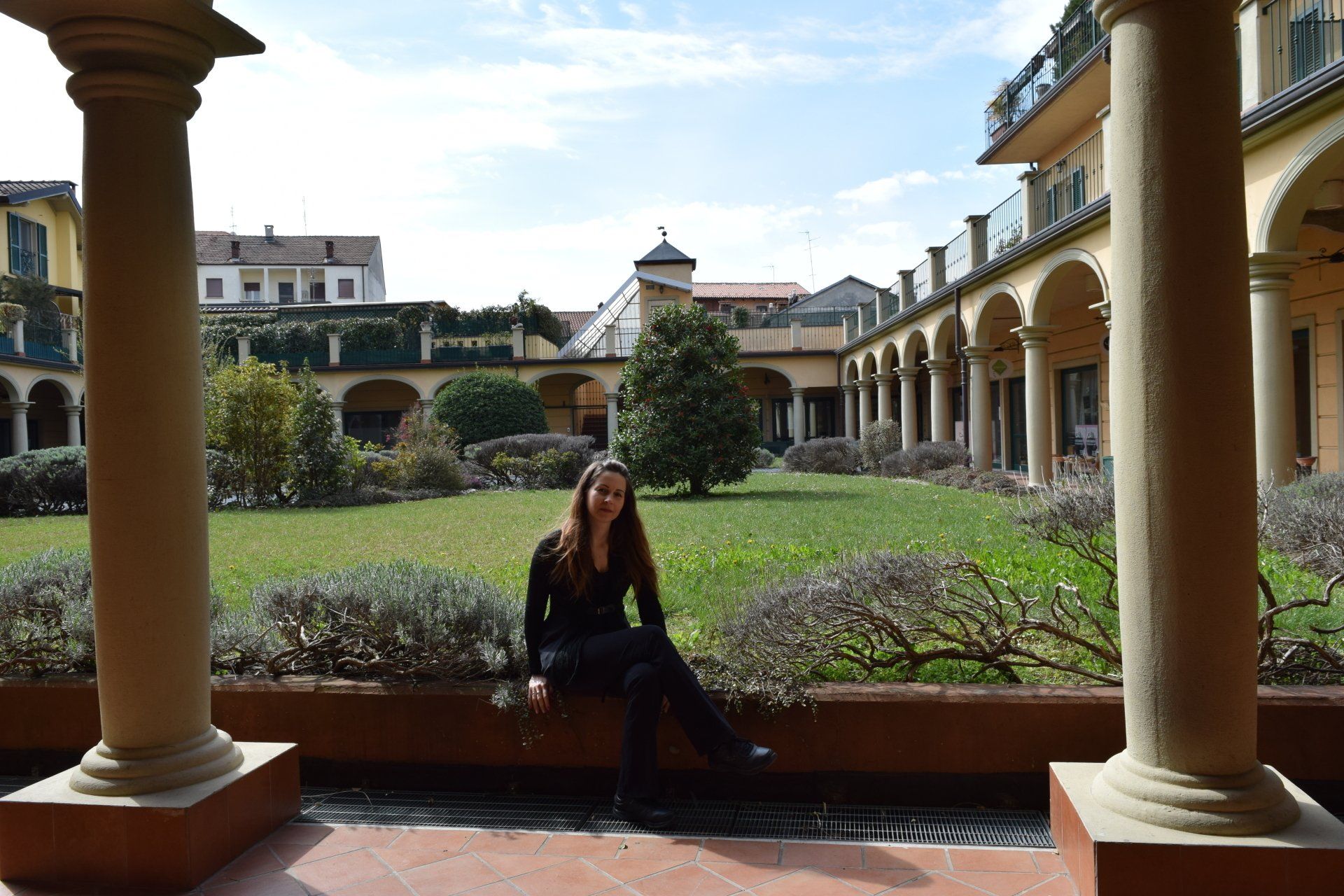 Una donna è seduta su un muretto basso tra colonne, con vista su un cortile con alberi e passaggi ad arco.