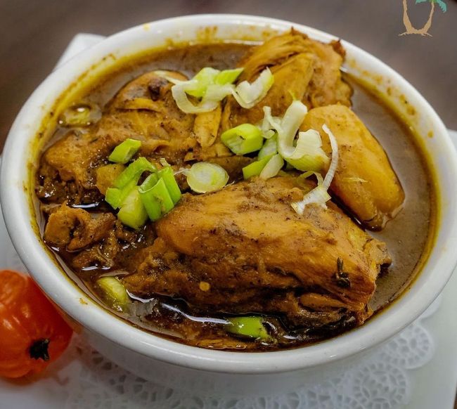 Bowl of Jamaican brown stew chicken, garnished with green onions, beside a whole scotch bonnet pepper.