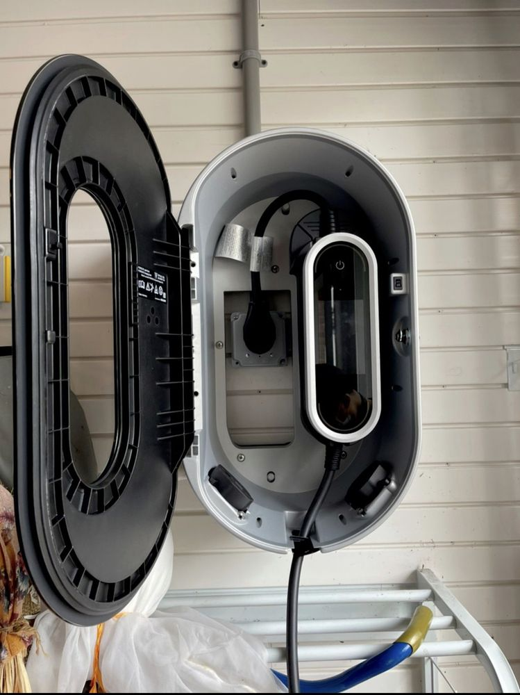 Open electric vehicle charger, white with black door, mounted on a wall. Charging cable hangs down.