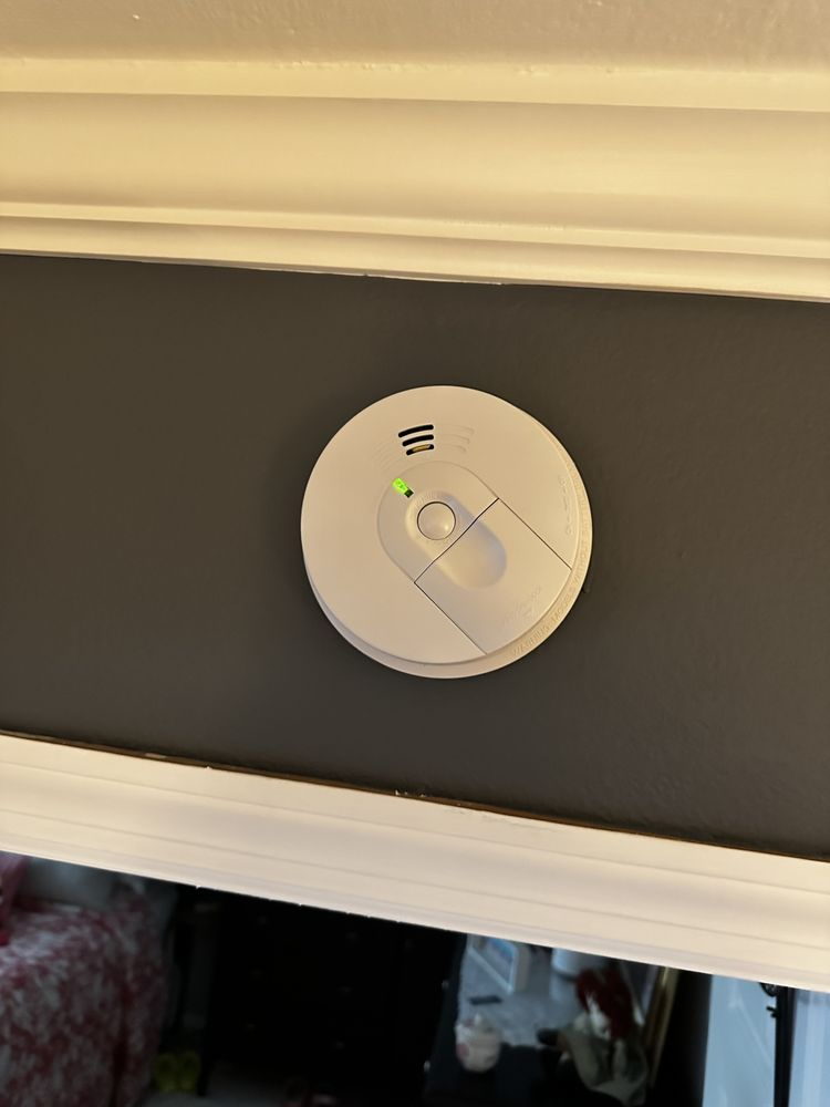 White smoke detector on a gray wall, near white trim; green light is on.