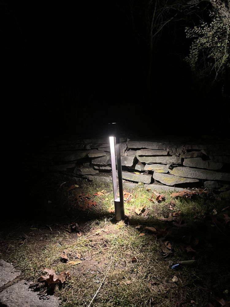 Lit outdoor pathway light with dark background; stone wall and grass visible.