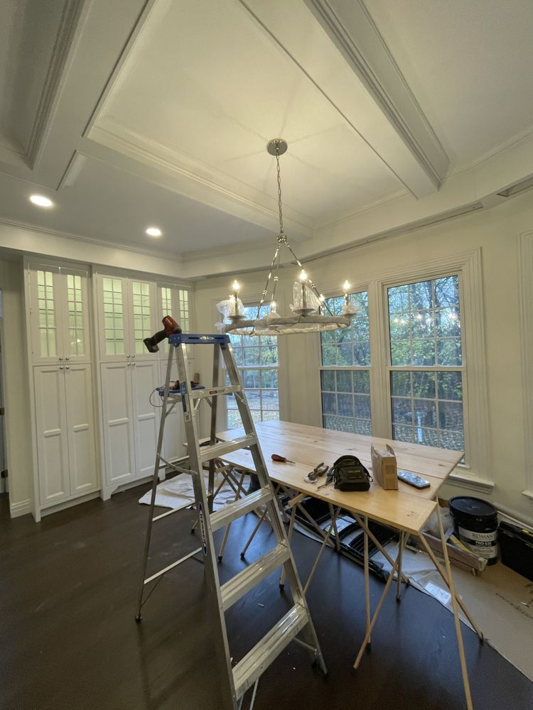 Room interior with ladder, chandelier, and unfinished table. Cabinets on left, windows on right.