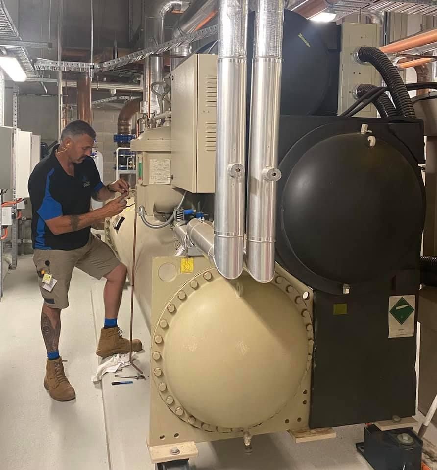 A man in work clothes repairs a large industrial chiller unit in a mechanical room. — Leader Air Conditioning in Albury/Wodonga, VIC