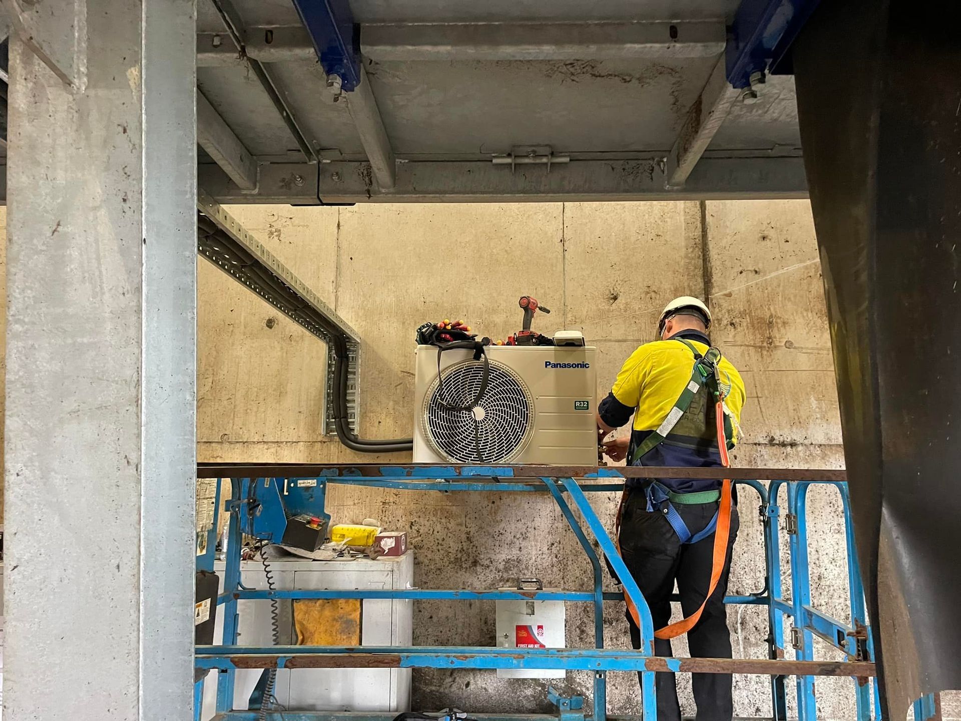A worker in a safety harness on a lift installs equipment near wiring in a factory setting. — Leader Air Conditioning in Queanbeyan, NSW