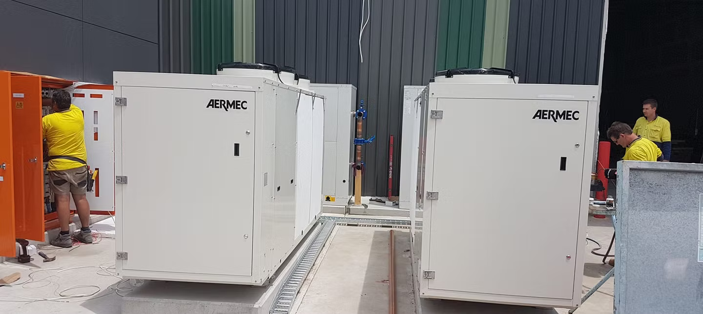 Workers installing industrial white machines in a warehouse. Orange and gray panels. — Leader Air Conditioning in Queanbeyan, NSW