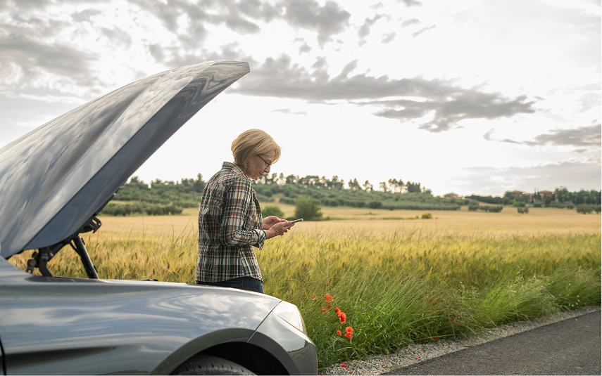 Woman with car trouble on a country road, checking her phone under the open hood, wheat field in background. | Jeremi's Auto Repair