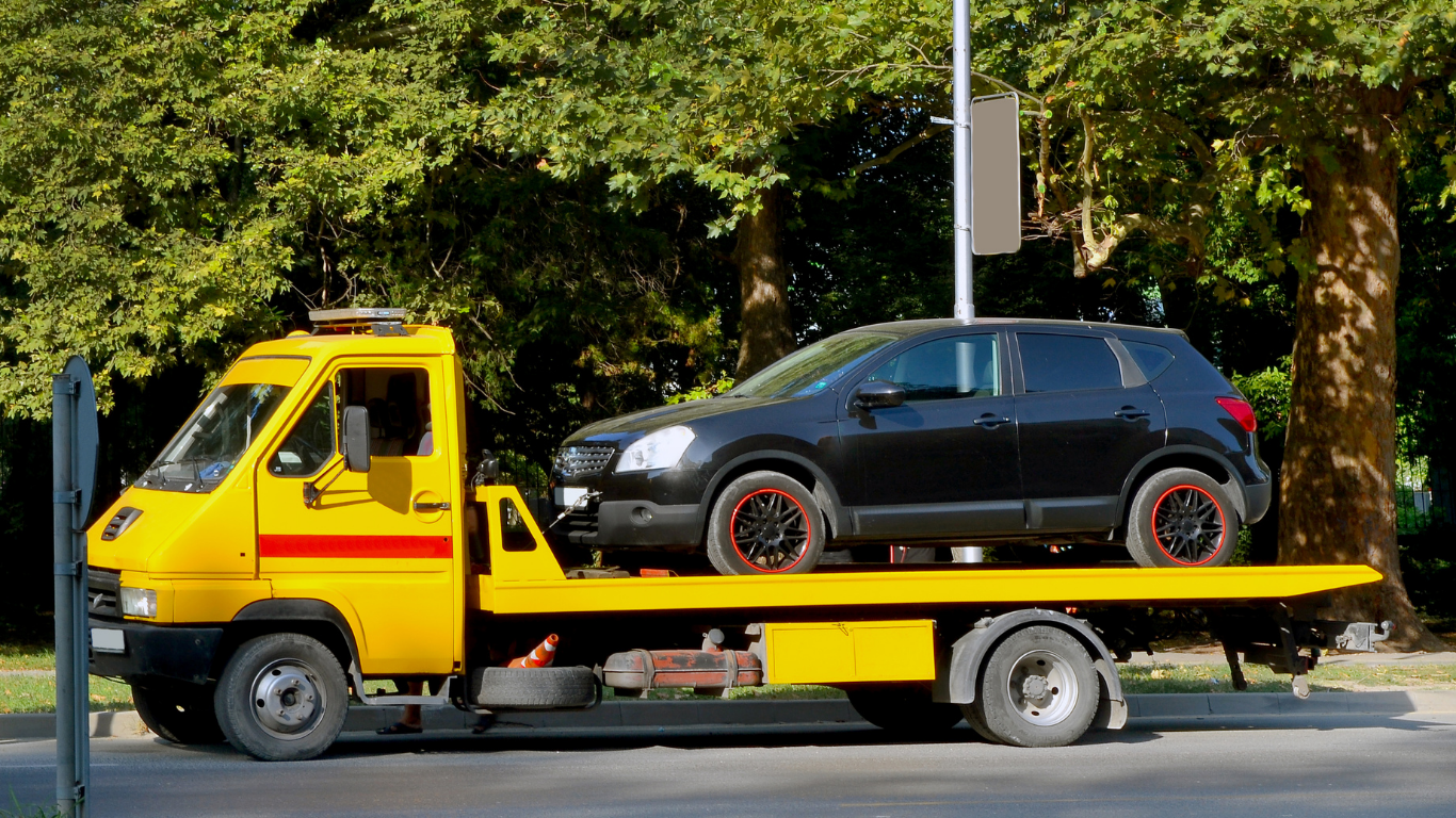 Yellow tow truck carrying a black SUV on a flatbed, parked by trees on a street.