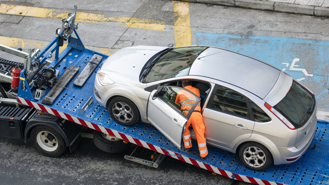 Silver car on blue tow truck platform, driver door open, worker in orange suit nearby