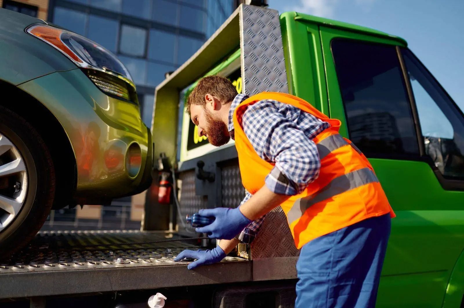 Tow truck operator in orange vest securing a car onto a green tow truck at the roadside