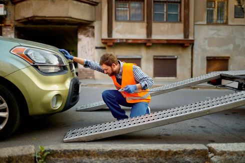 Tow truck operator loading a green car onto a flatbed ramp on a city street