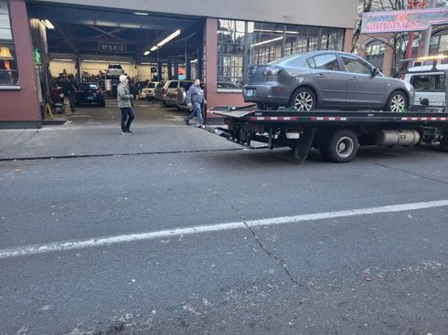 Car carrier trailer with a gray sedan outside a shop, with a few people in the background.