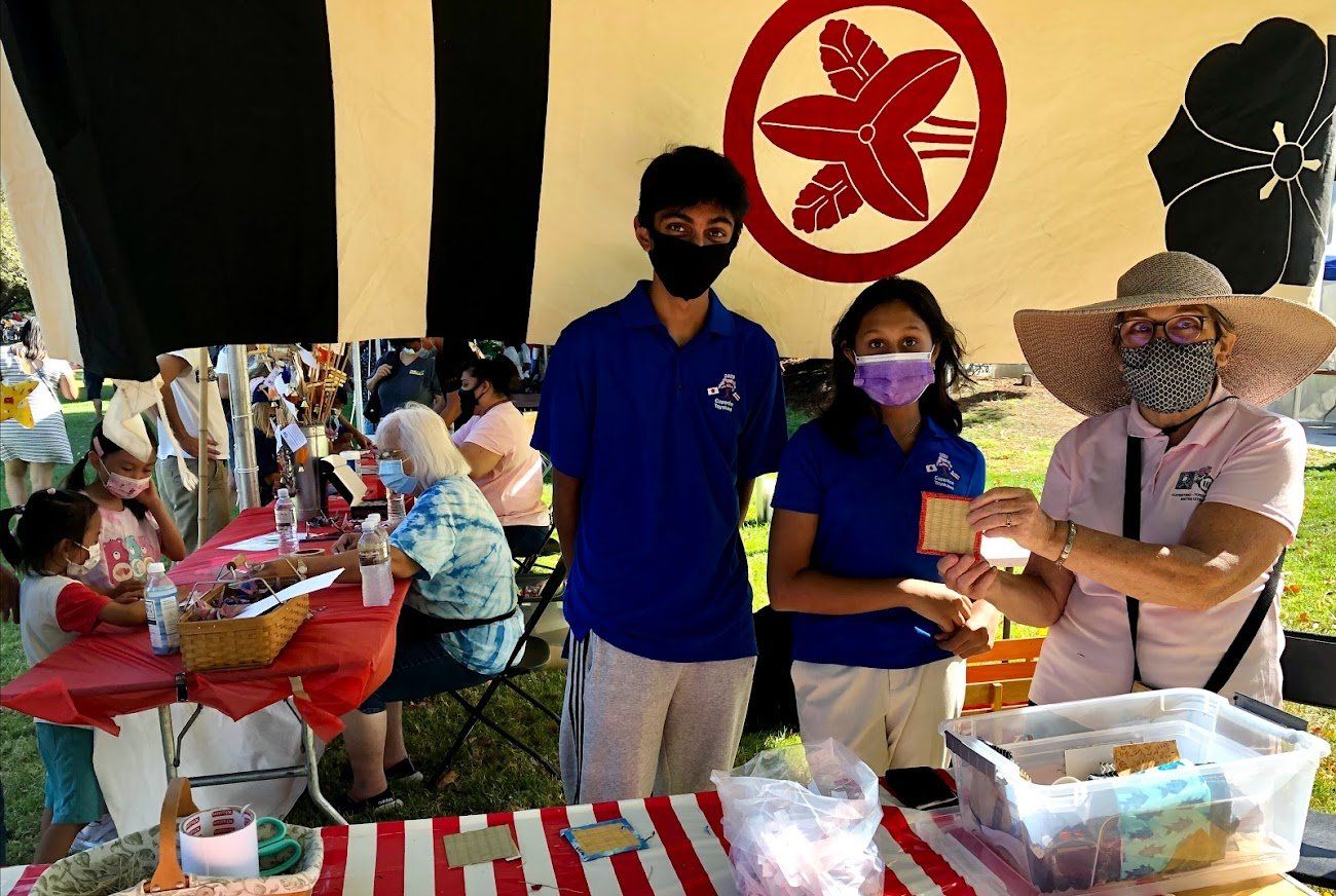 People behind a red-and-white checkered booth at an indoor market or fair, wearing masks and a straw hat.