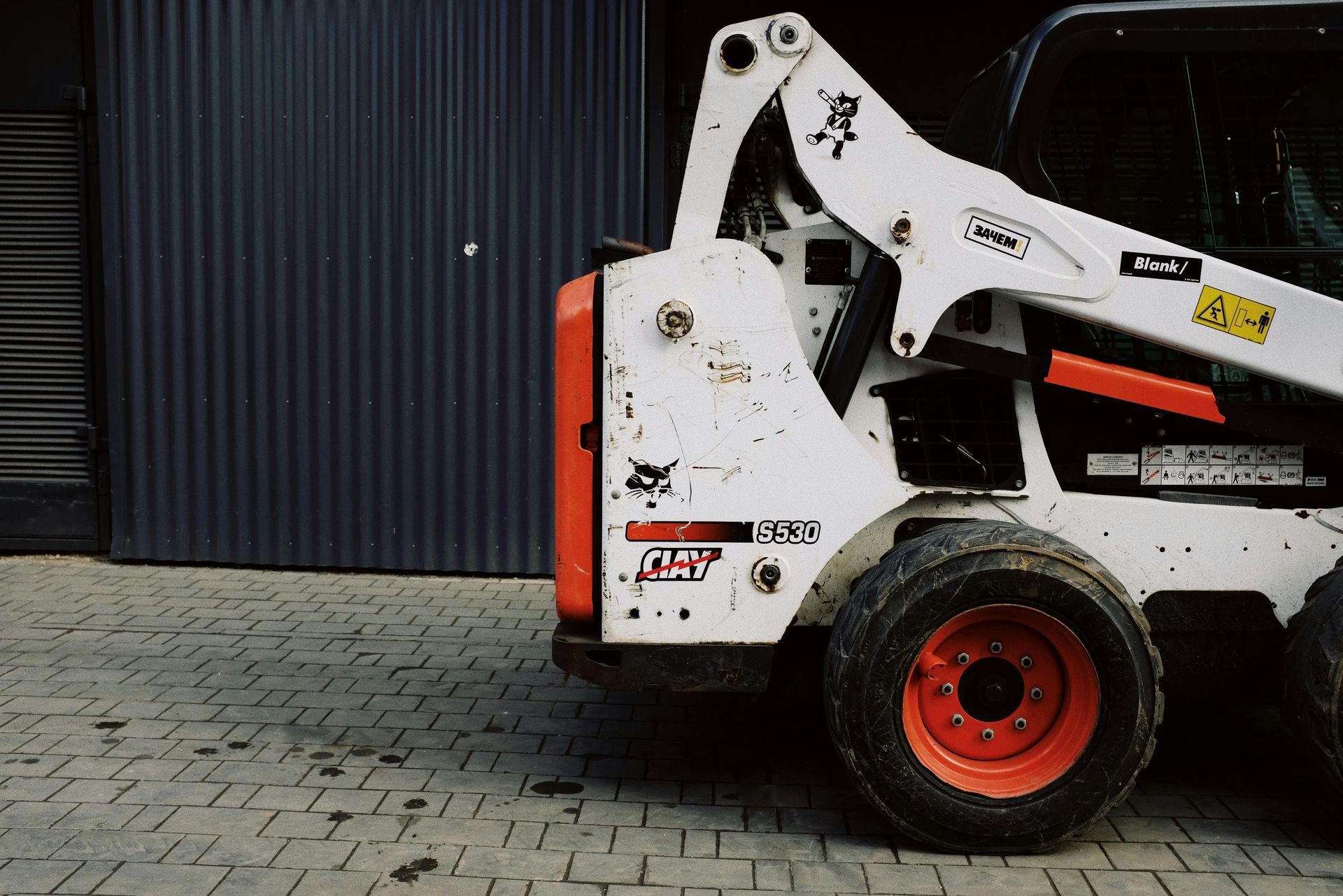White skid-steer loader with orange wheels parked beside a dark corrugated wall on brick pavement